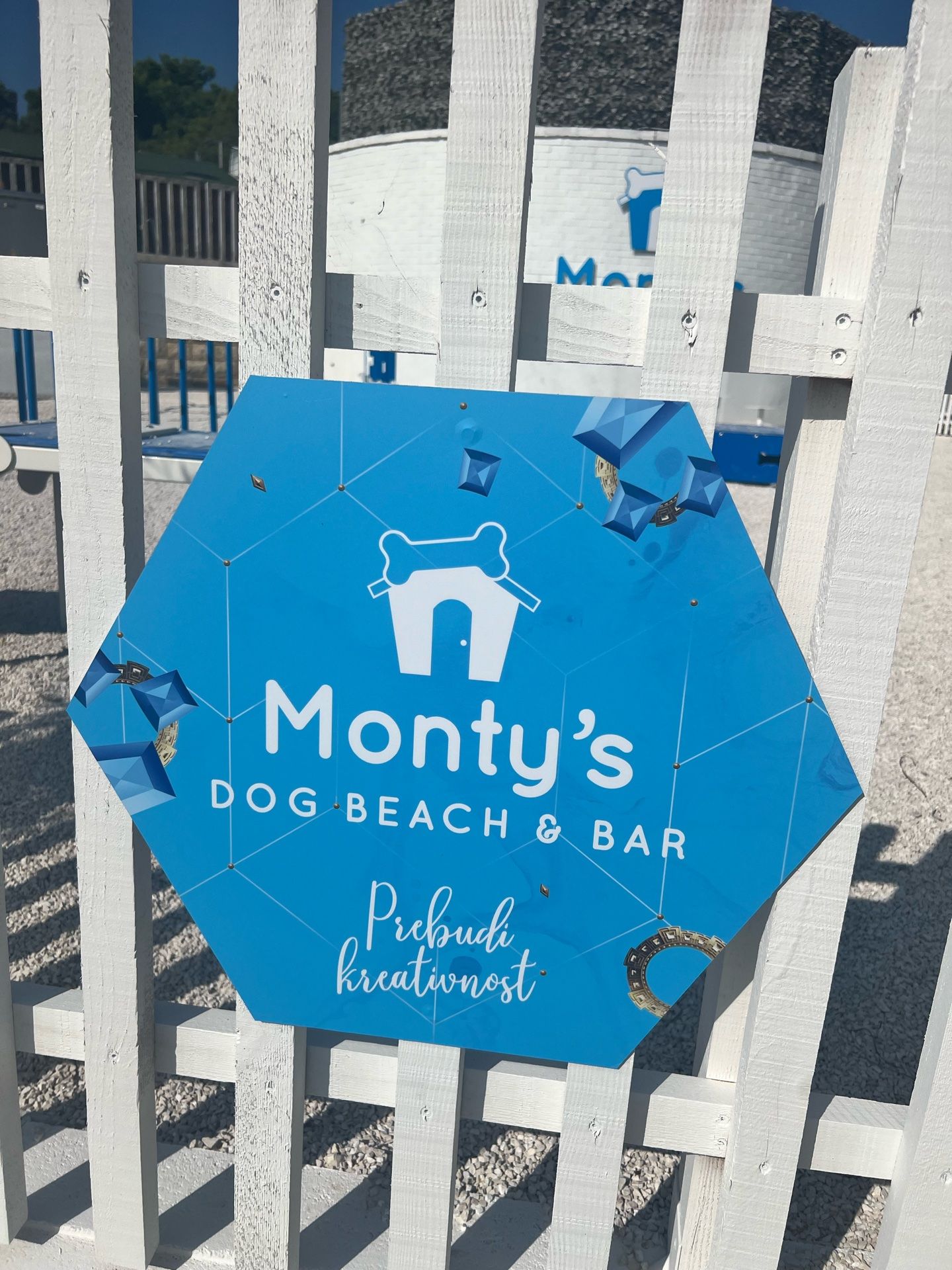 Ein blauer, sechseckiger Schild mit der Aufschrift 'Monty's Dog Beach & Bar' vor einem weißen Zaun.