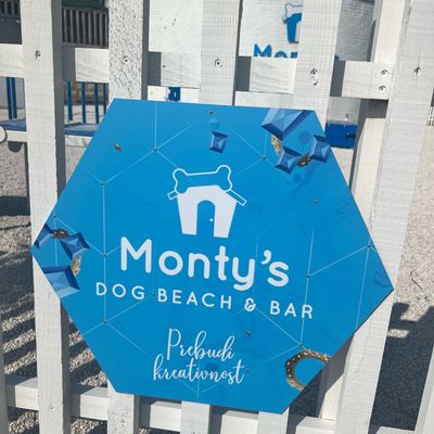 Ein blauer, sechseckiger Schild mit der Aufschrift 'Monty's Dog Beach & Bar' vor einem weißen Zaun.
