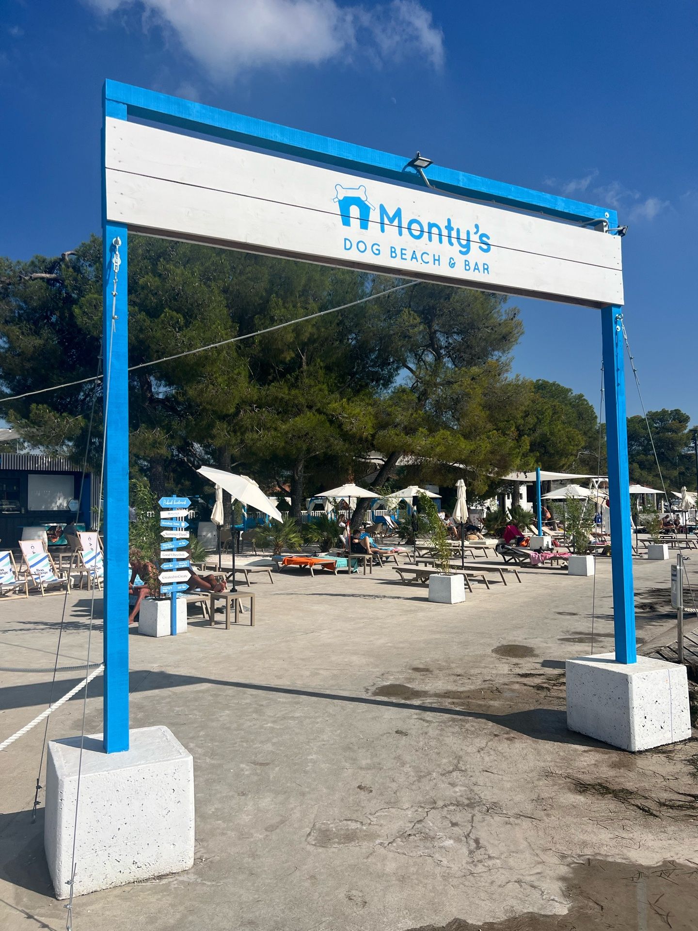 Blaues Eingangsschild von Monty's Dog Beach & Bar mit Liegen und Sonnenschirmen im Hintergrund.