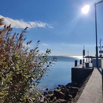 Blick auf eine steinige Küste und einen Hafen bei Sonnenschein mit blauem Himmel und wenigen Wolken.