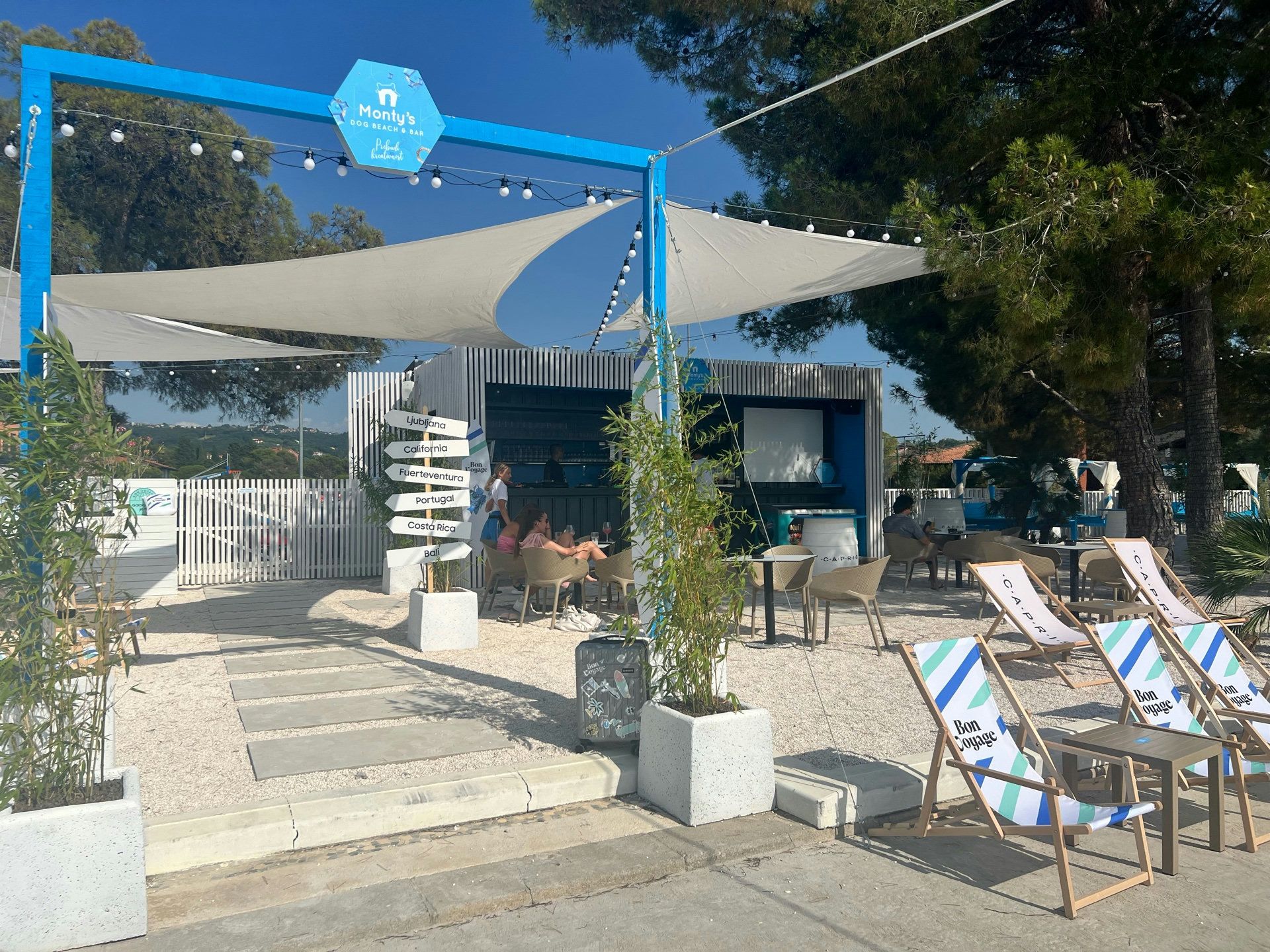 Außenbereich einer Hundestrandbar mit blauem Rahmen, Sonnensegeln, Sitzgruppen und einem Wegweiser.