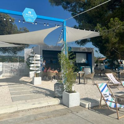 Außenbereich einer Hundestrandbar mit blauem Rahmen, Sonnensegeln, Sitzgruppen und einem Wegweiser.