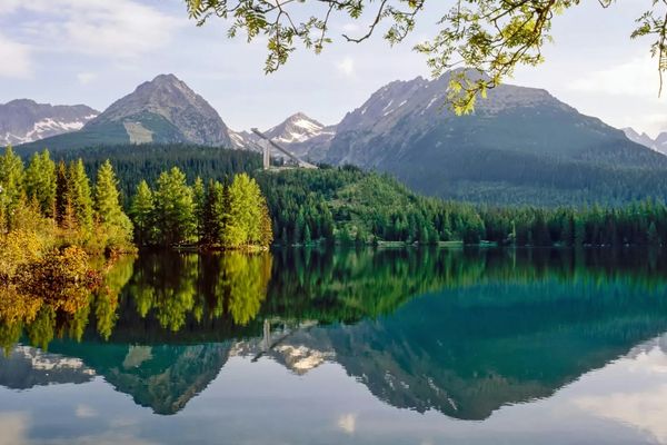 Klarer Bergsee mit Baumreflexionen, umgeben von bewaldeten Bergen.